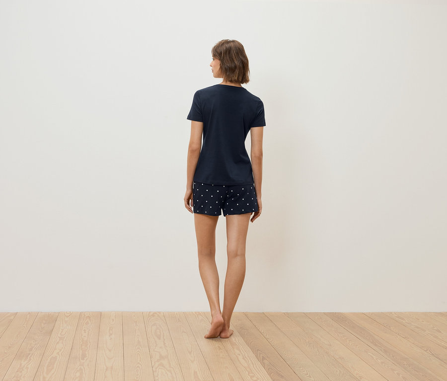 Egy nő háttal áll a kamerának, sötétkék pólót és fehér pöttyös rövidnadrágot visel.