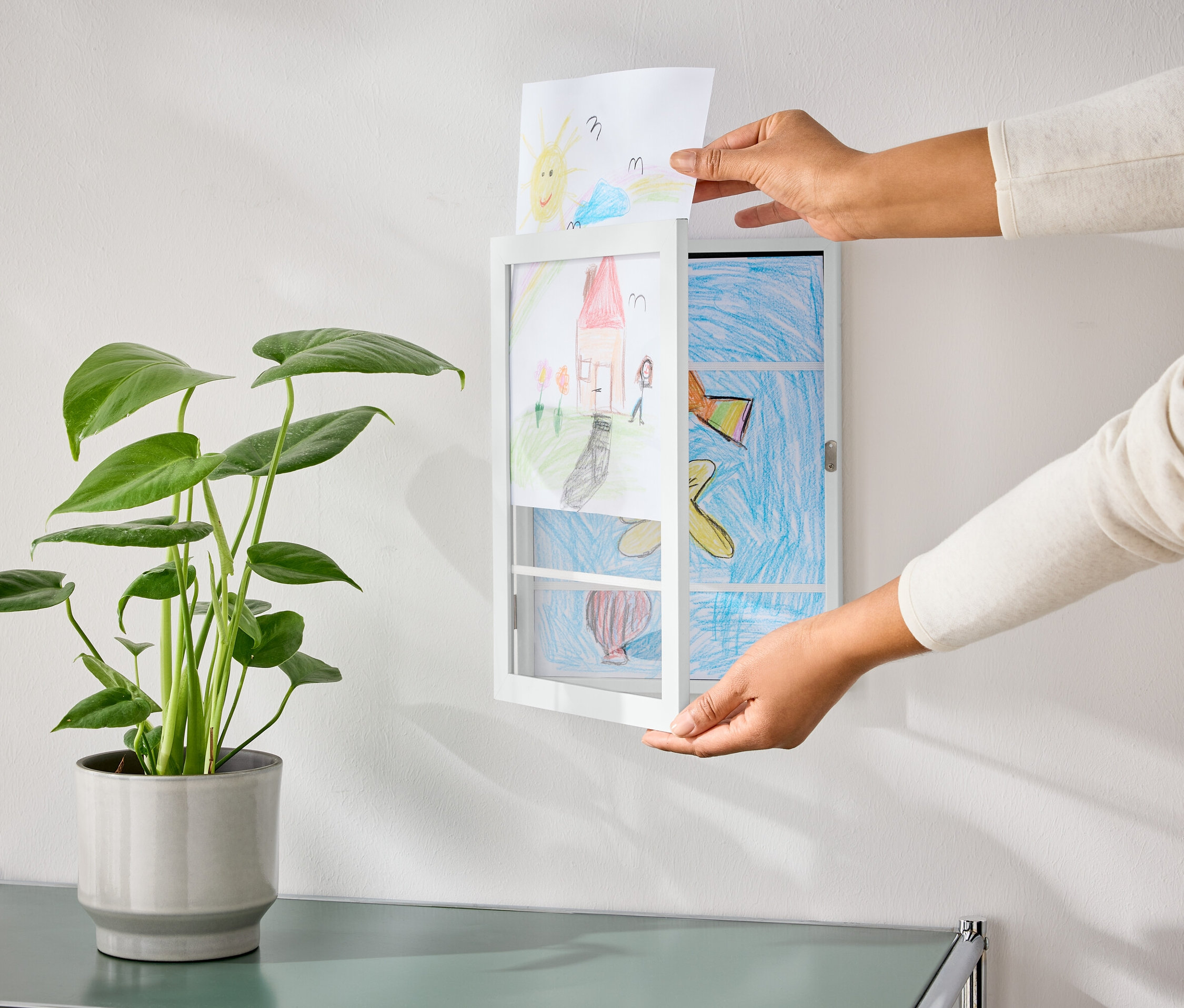 Kezek egy rajzot helyeznek egy felnyitható képkeretbe, amely egy cserepes növény mellett lóg a falon.