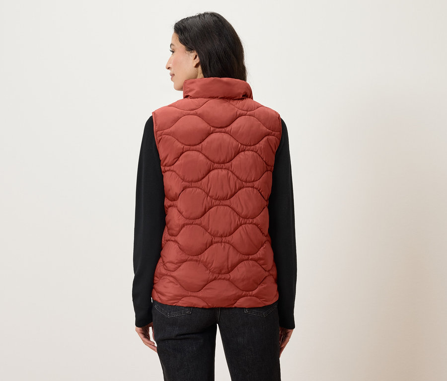 A nő háttal áll a kamerának, narancssárga, steppelt mellényt és fekete, hosszú ujjú inget visel.