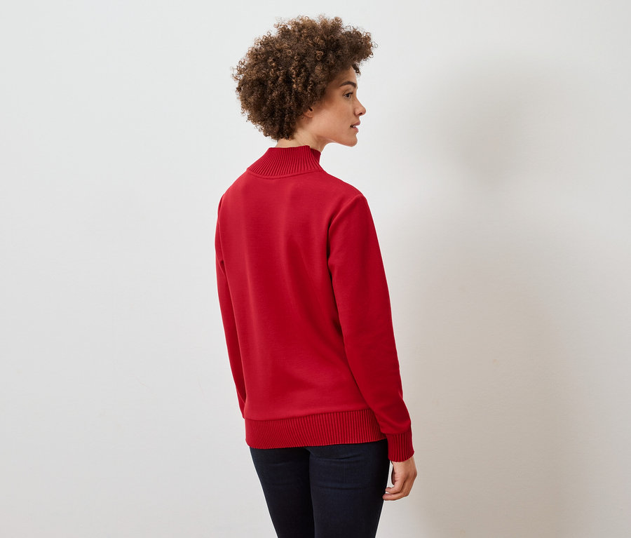 Göndör hajú nő áll oldalt, piros pulóvert és sötétkék farmert visel.