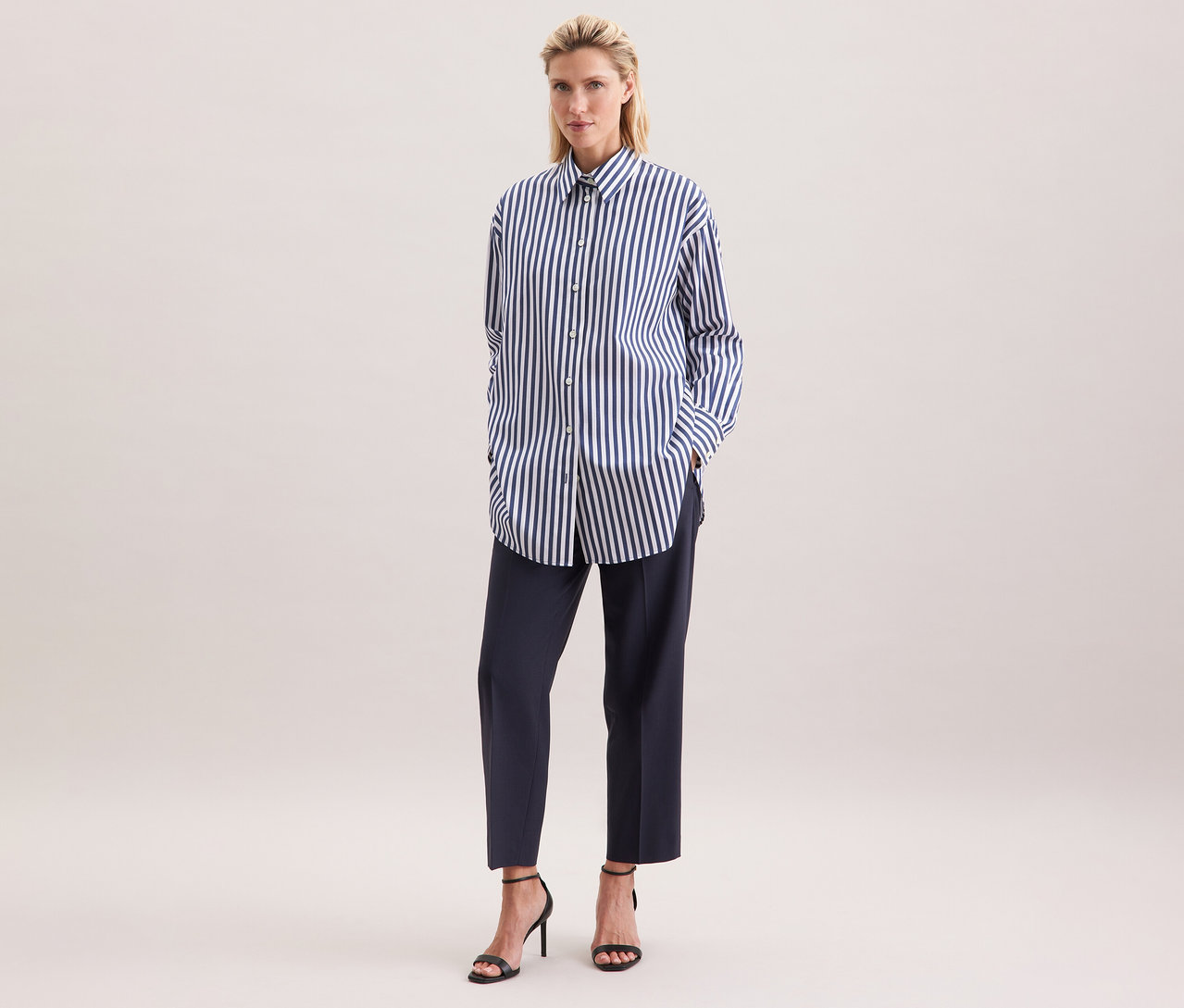 Nő visel egy csíkos Seidensticker túlméretes hosszú ingblúzt, sötét nadrágot és fekete magassarkút.