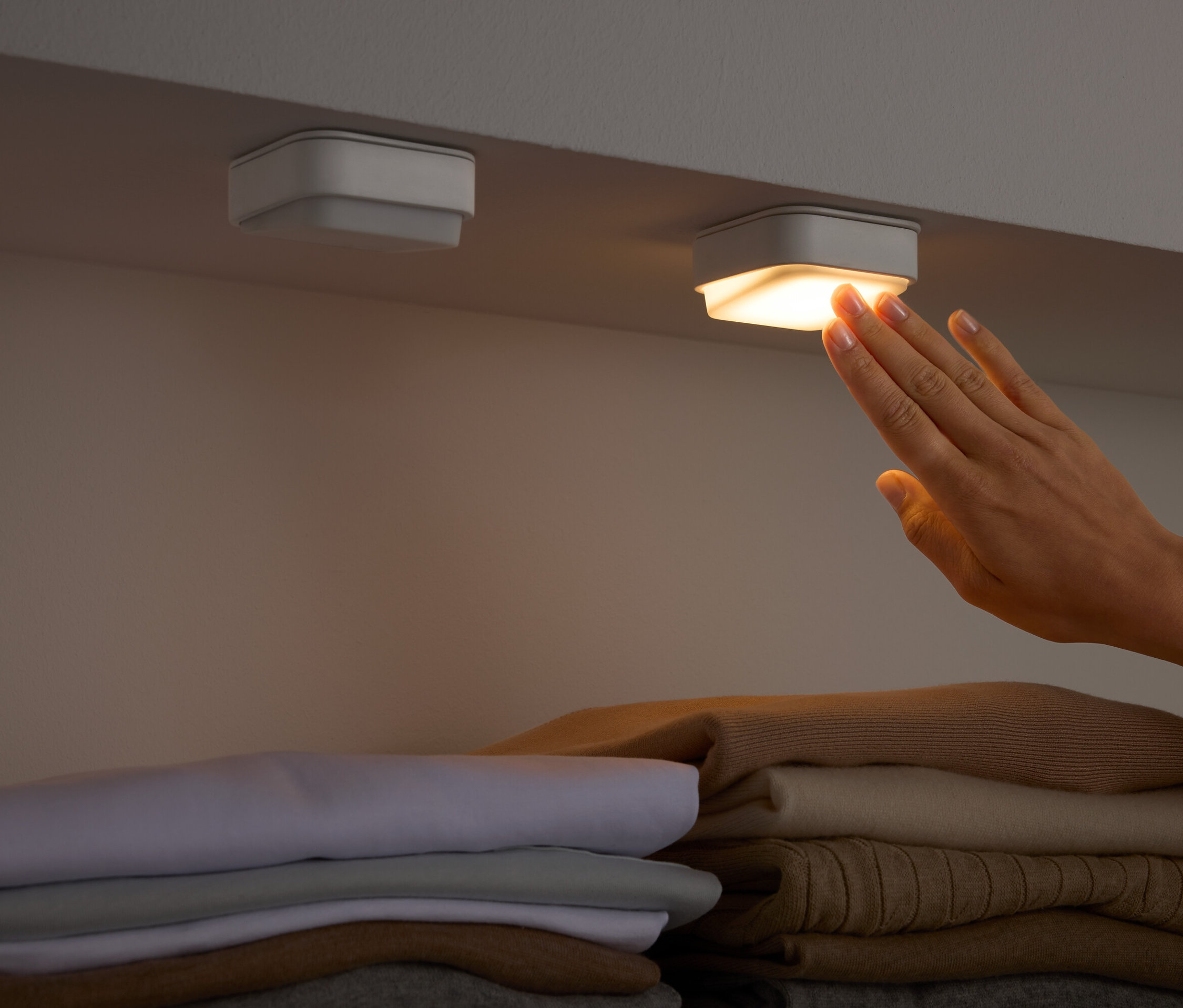 Egy kéz megérint egy négyzet alakú lámpát egy polc alatt, összehajtott ruhák felett.