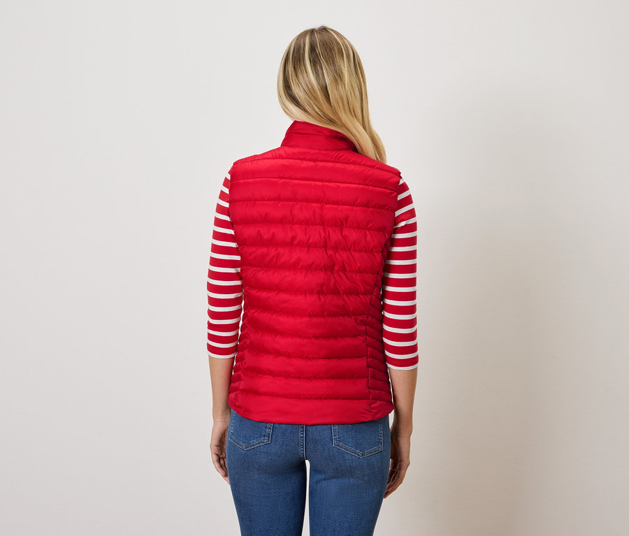 Hátulról látható egy szőke hajú nő, aki piros steppelt mellényt és kék farmert visel.