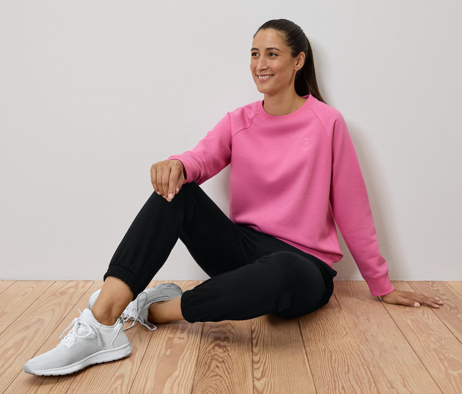 Nő ül a fapadlón, rózsaszín pulóvert, fekete nadrágot és fehér tornacipőt visel.
