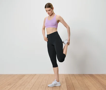 Egy nő nyújtja a lábát, miközben a bokáját tartja a kezében. Lila sportmelltartót, fekete leggingset és fehér tornacipőt visel.