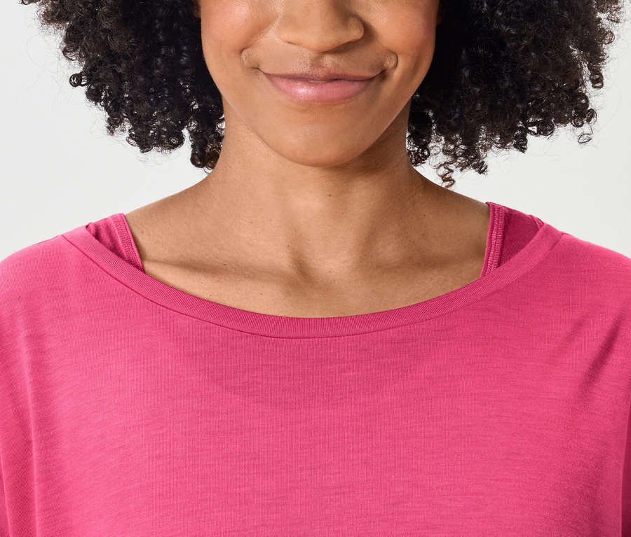 Egy mosolygó, sötét, göndör hajú nő felsőteste rózsaszín pólóban.