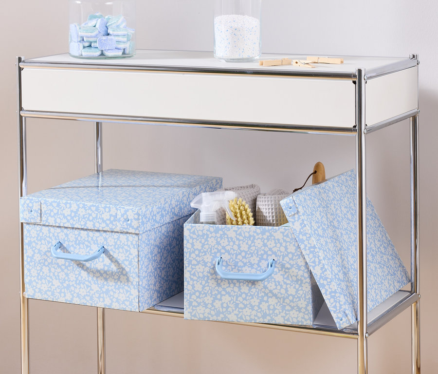 Egy polcon két kék tárolódoboz fedővel és egy tárolódoboz fedővel, benne fehér törölközőkkel és egy kefével.