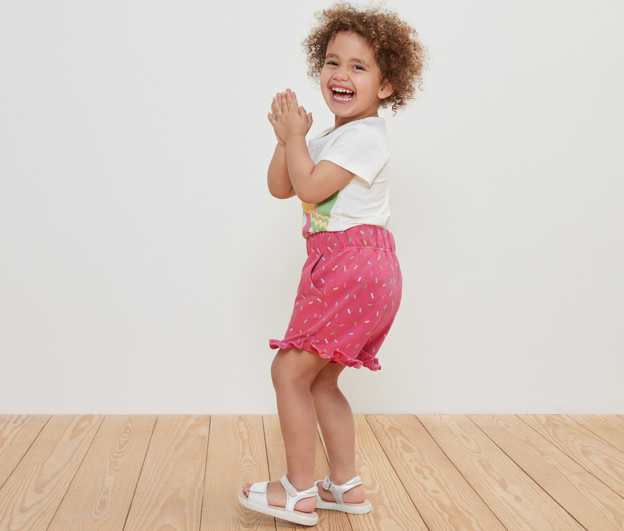 Mosolygó, göndör hajú kislány tapsol szabadidős rövidnadrágban.