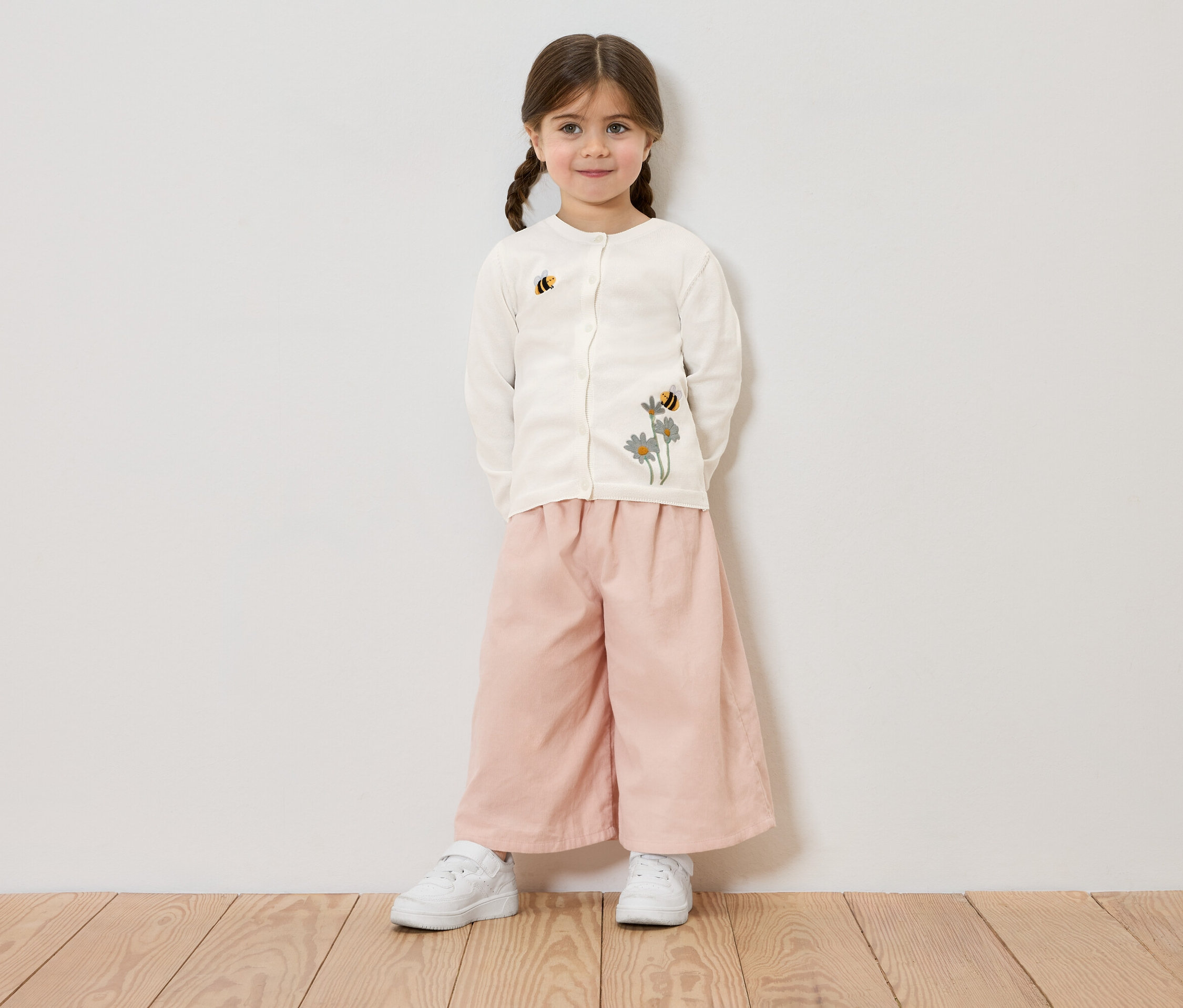Lány copfokkal, virág- és méhimintás krémszínű kardigánt, rózsaszín nadrágot és fehér tornacipőt visel, fehér fal előtt.
