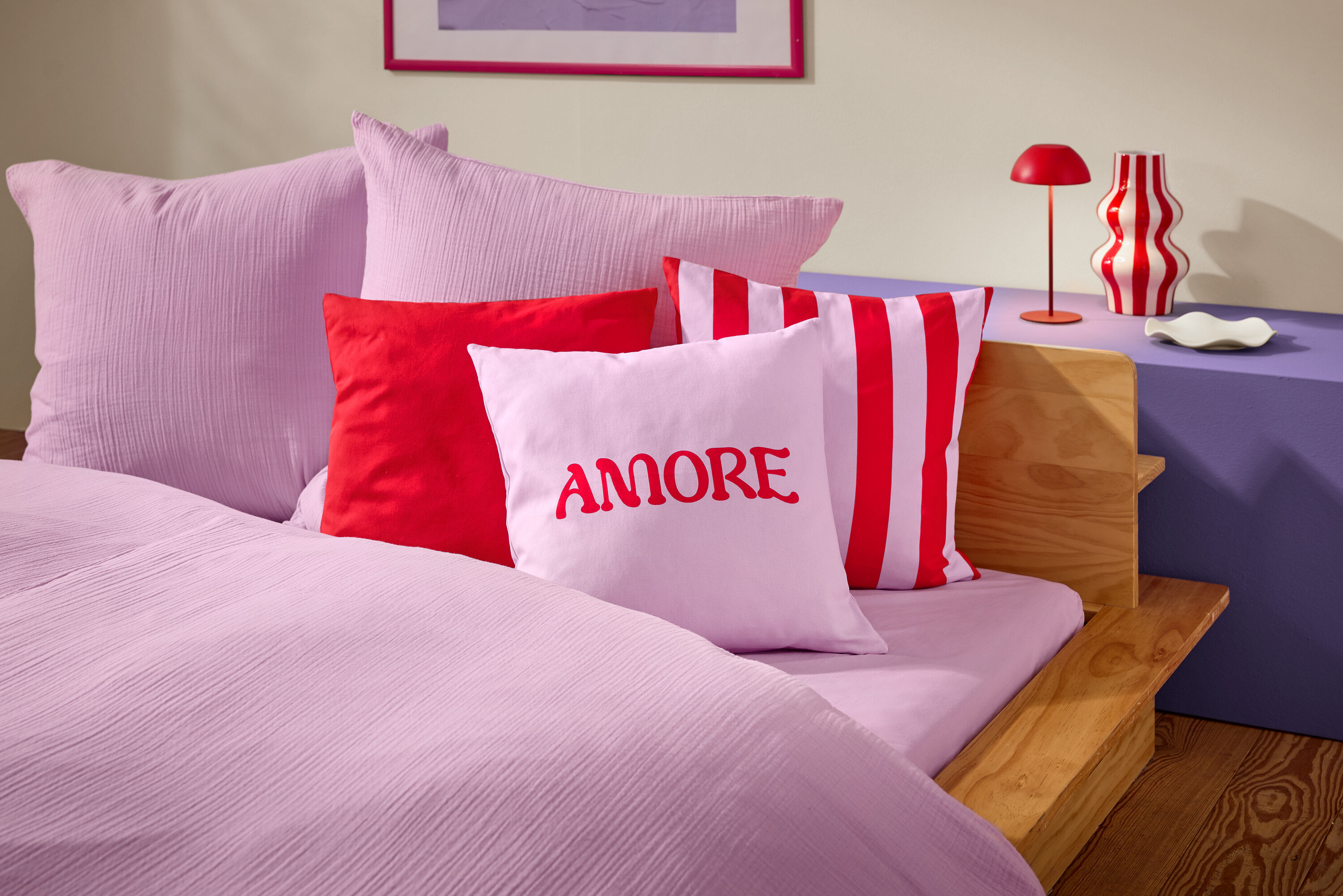 Egy ágy közeli képe rózsaszín ágyneművel és párnákkal. Az egyik párnán piros betűkkel az AMORE szó látható.