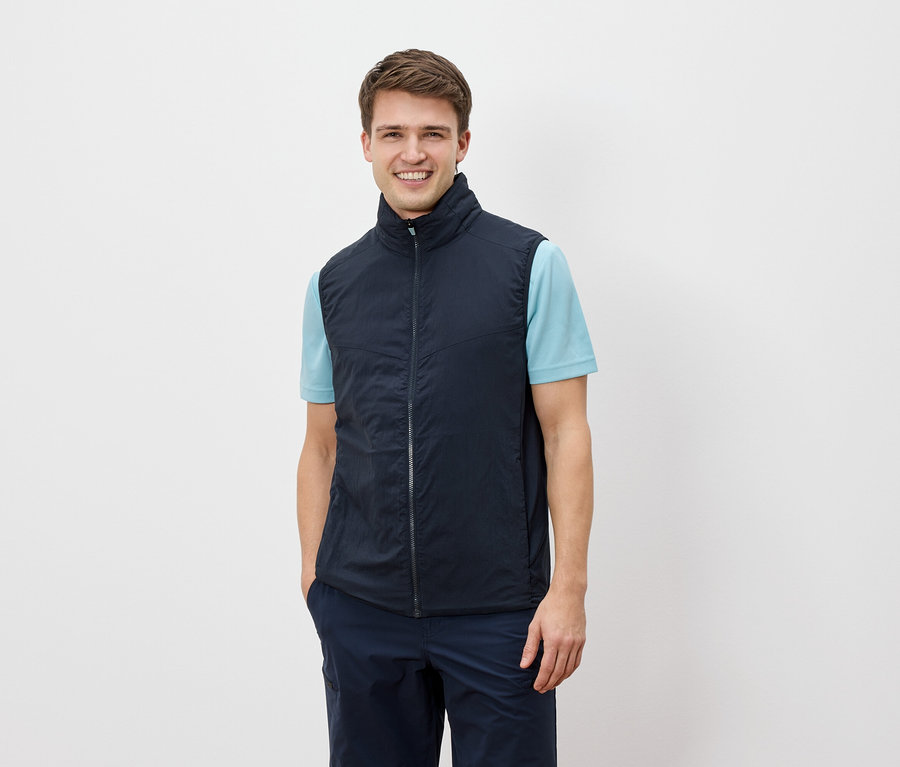 Egy férfi mosolyog a kamerába, és egy tengerészkék mellényt visel egy világoskék póló felett.