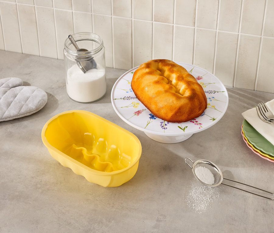 A konyhapulton egy virágos állványon kuglóf, egy sárga kuglófforma, egy üveg cukor, egy szita és edényfogó található.