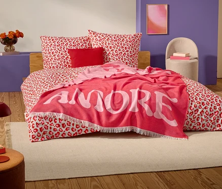 Egy vörös-fehér leopárdmintás ágyon egy piros takaró fekszik, nagy fehér betűkkel az "AMORE" szóval.