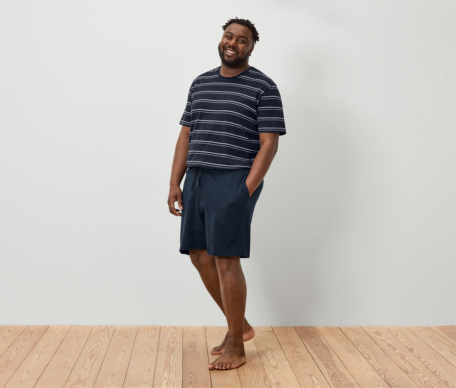 Egy mosolygó férfi áll mezítláb egy fehér fal előtt, csíkos pólót és sötétkék rövidnadrágot visel.