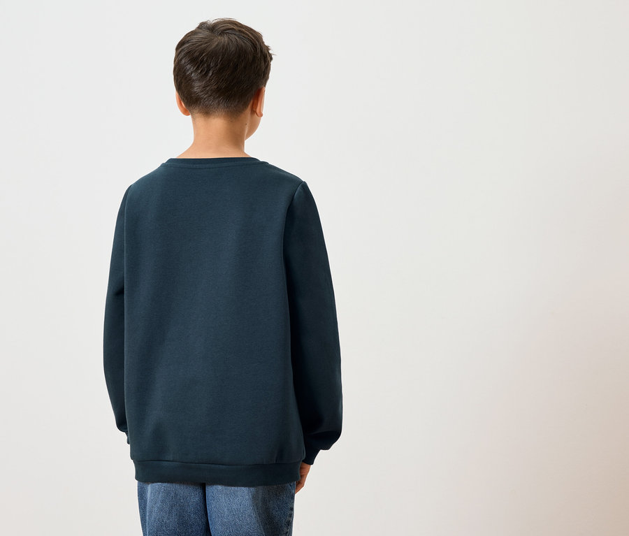 Hátulnézet egy sötét hajú gyermekről, aki sötétkék pulóvert és kék farmert visel fehér háttér előtt.