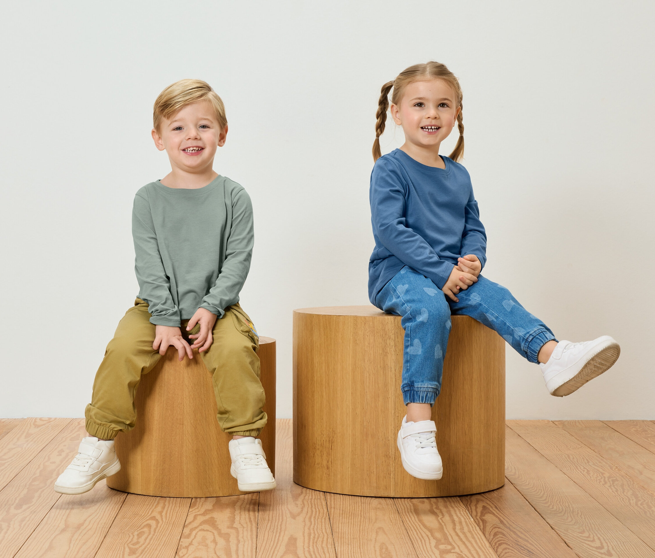 Egy fiú zöld hosszú ujjú pólóban és khaki nadrágban ül egy kislány mellett, aki kék hosszú ujjú pólót és szívecskés farmernadrágot visel, fa székeken.