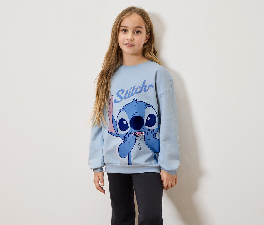 Hosszú szőke hajú lány világoskék Stitch mintás pulóvert és fekete leggingset visel.