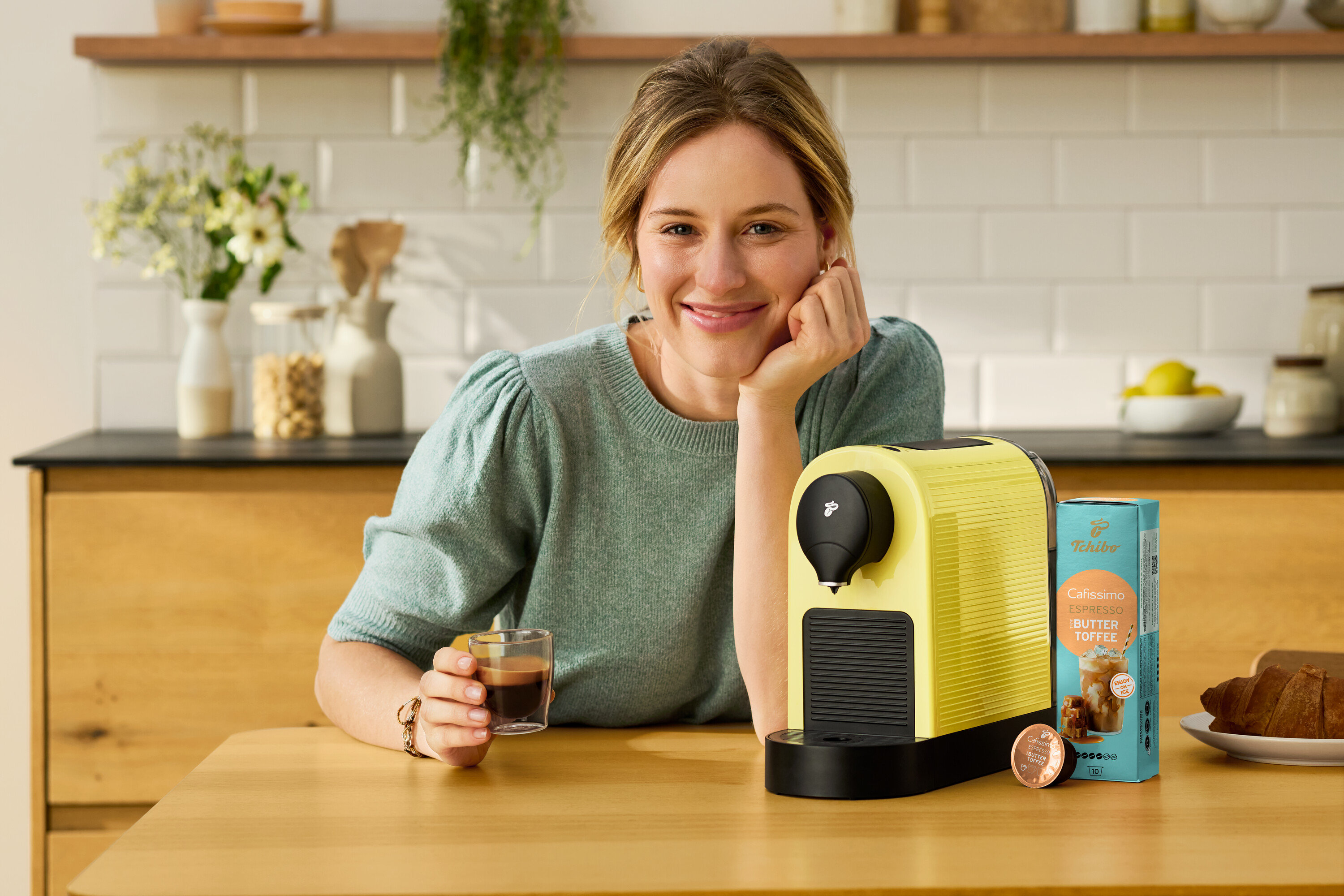 Nő ül az asztalnál egy csésze kávéval, egy sárga Cafissimo pure+ kávéfőzővel és egy Cafissimo Flavoured Espresso – Toasted Nut – 10 kapszula csomaggal.