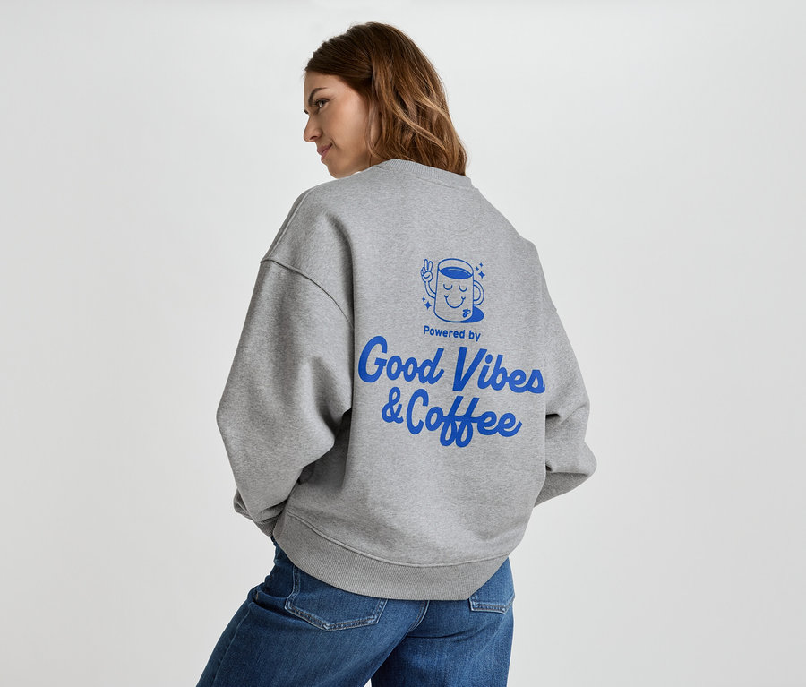 Nő szürke pulóvert visel, hátán kék "Good Vibes & Coffee" felirattal, hozzá farmernadrágot.