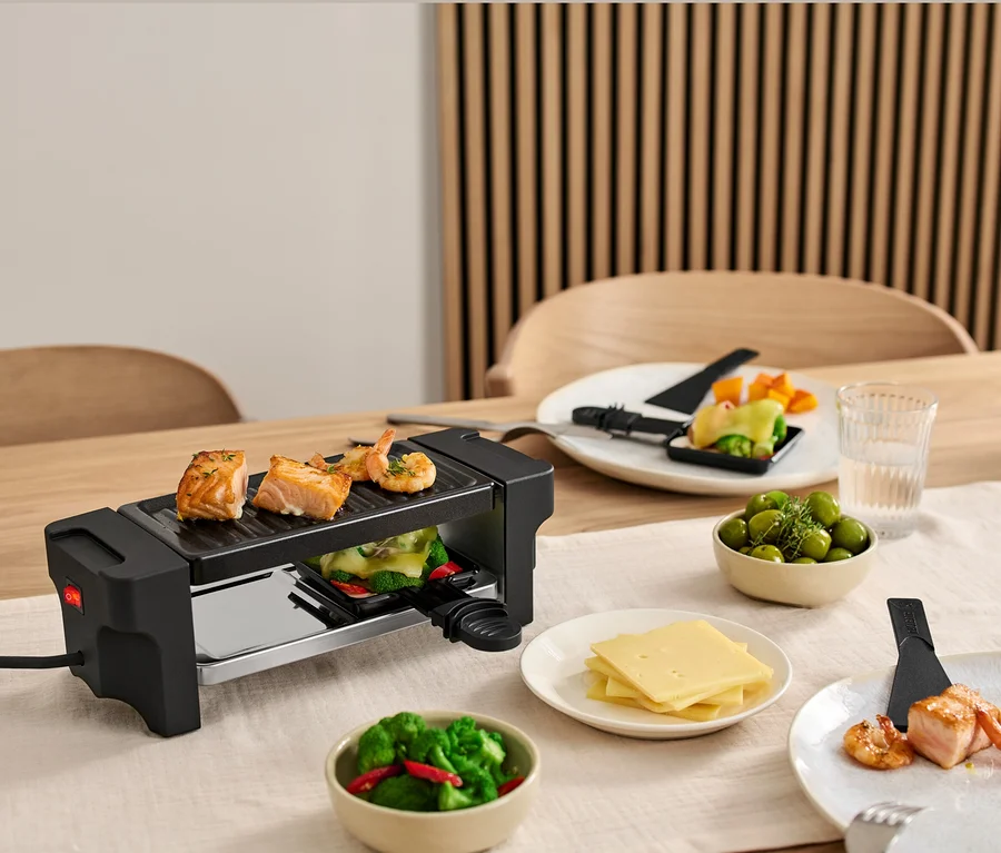 Egy kétszemélyes raclette grill van az asztalon, lazaccal és garnélával a grillen. Tányérok hozzávalókkal a grill körül.