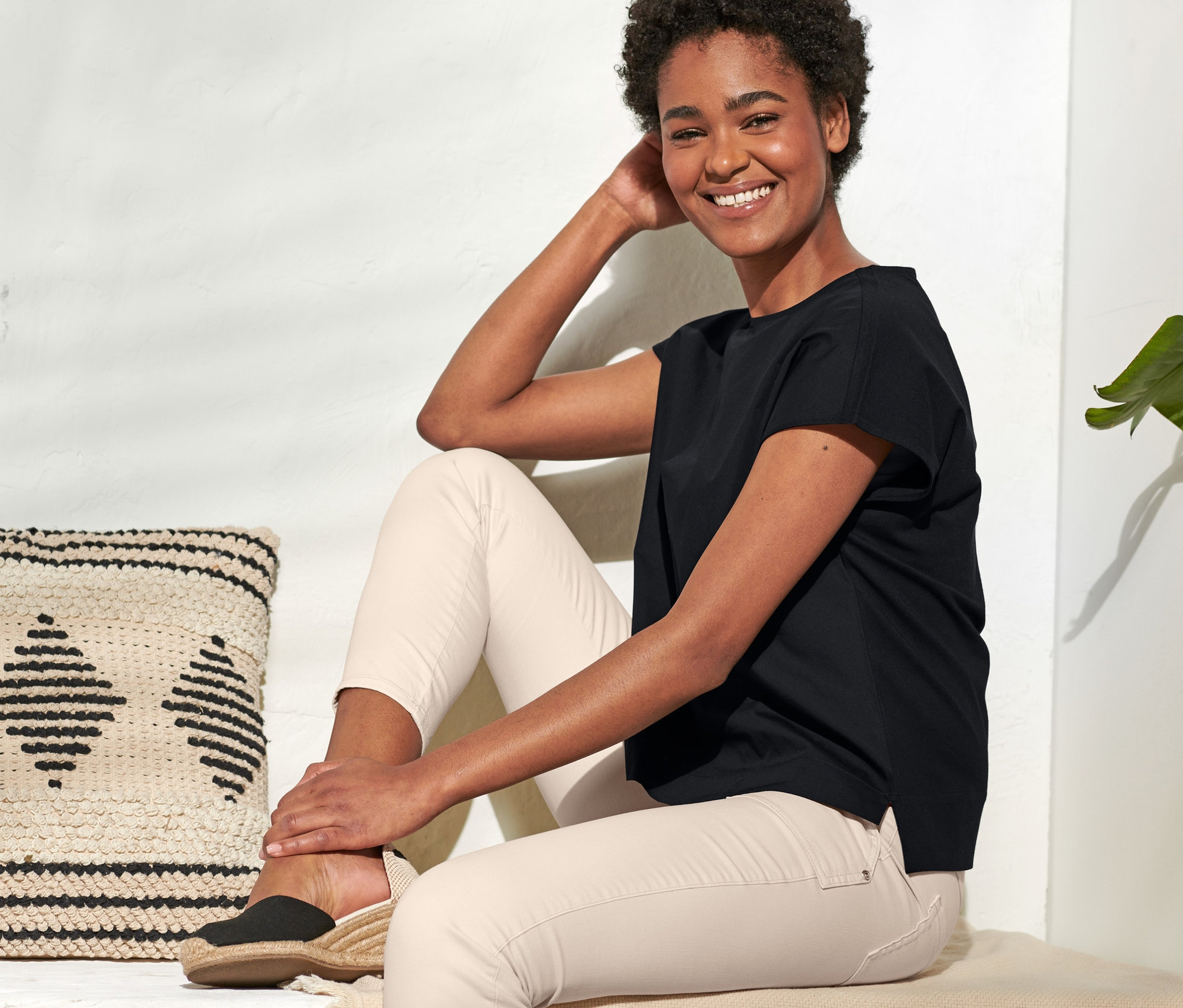 Egy mosolygó nő ül, fekete női pólóblúzt és világos nadrágot visel.