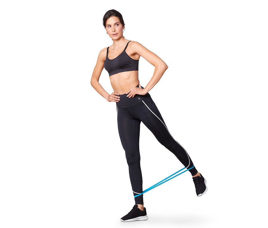 Nő edz kék fitneszszalaggal, fekete sportmelltartót és leggingst visel. A háttérben fehér háttér látható. Csípőre tett kézzel.