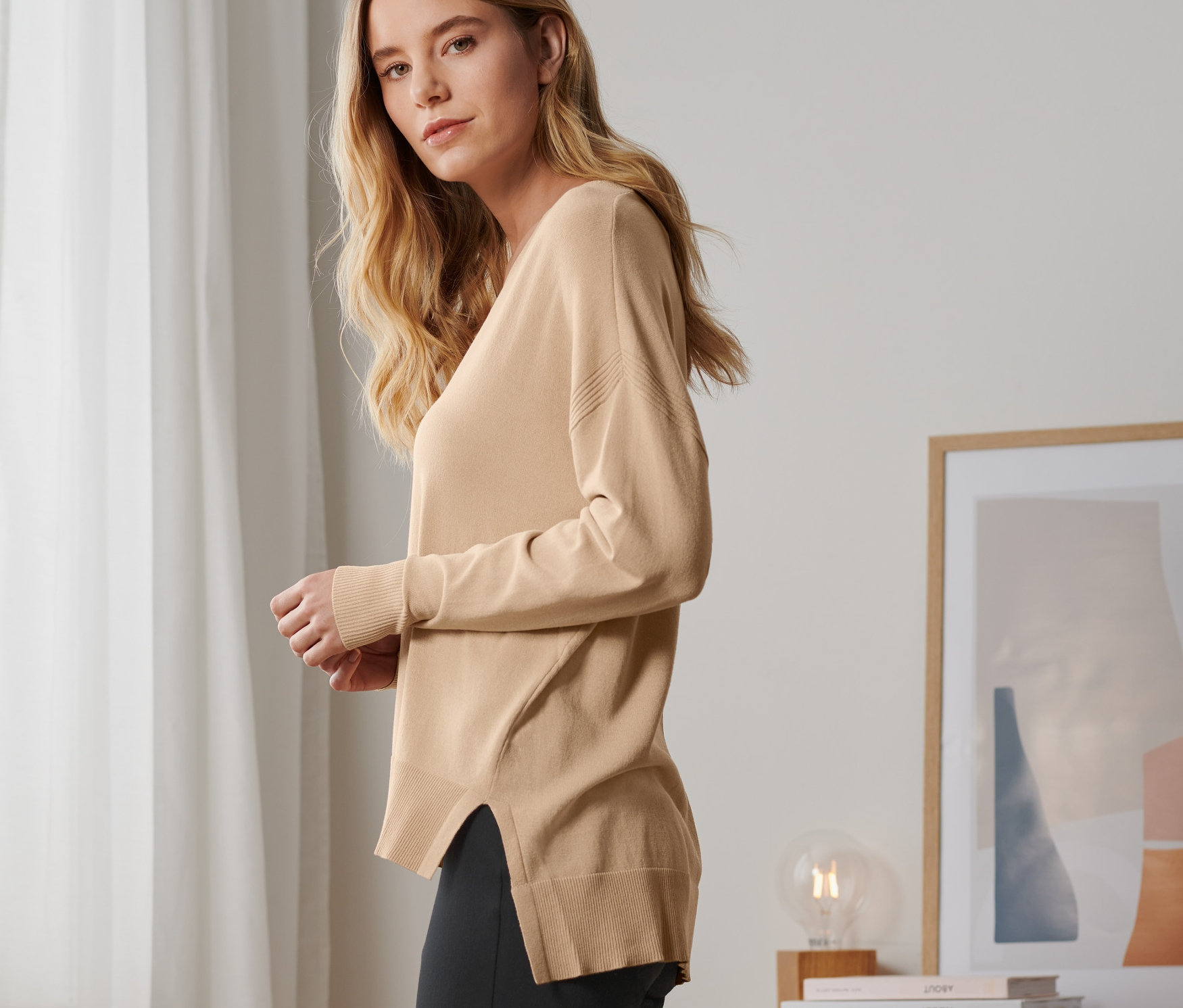 Egy szőke nő pózol egy bézs színű, finomkötésű pulóverben és fekete nadrágban.