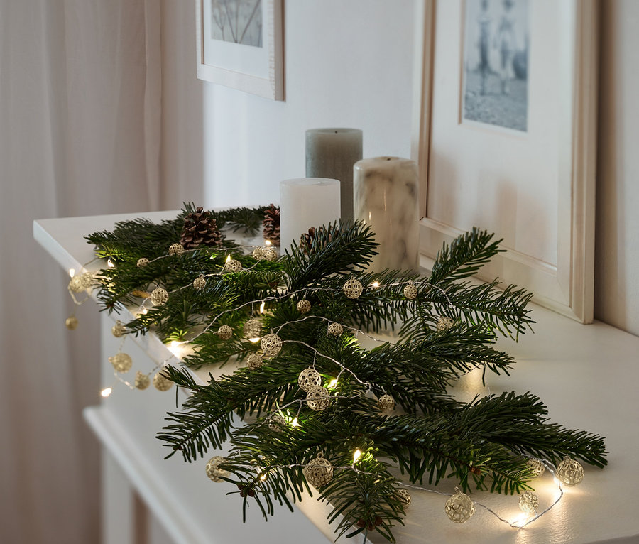 Fenyőágak és három gyertya fekszik egy kandallópárkányon két bekeretezett kép alatt. Az ágakon egy LED-es égősor, 38 gömb fekszik.