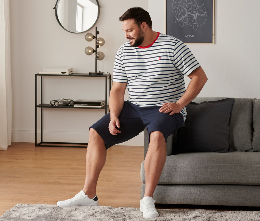 Férfi ül a kanapén, fehér sneaker cipőt, tengerészkék chino rövidnadrágot és egy csíkos férfi pólót (2 db-os szettből) visel.