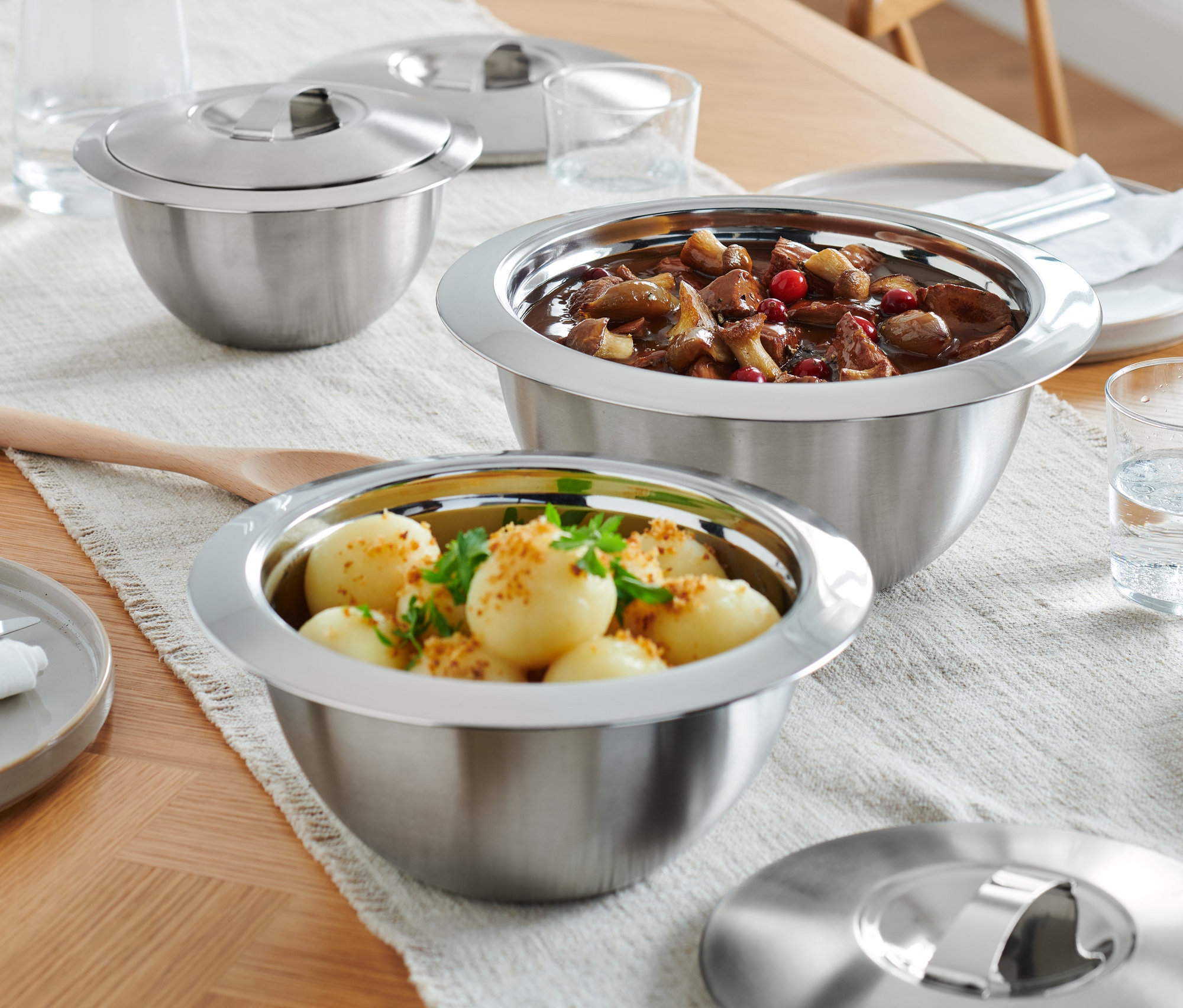 A faasztalon három tálalótál nemesacél fedővel, tányérok és vizespoharak vannak.