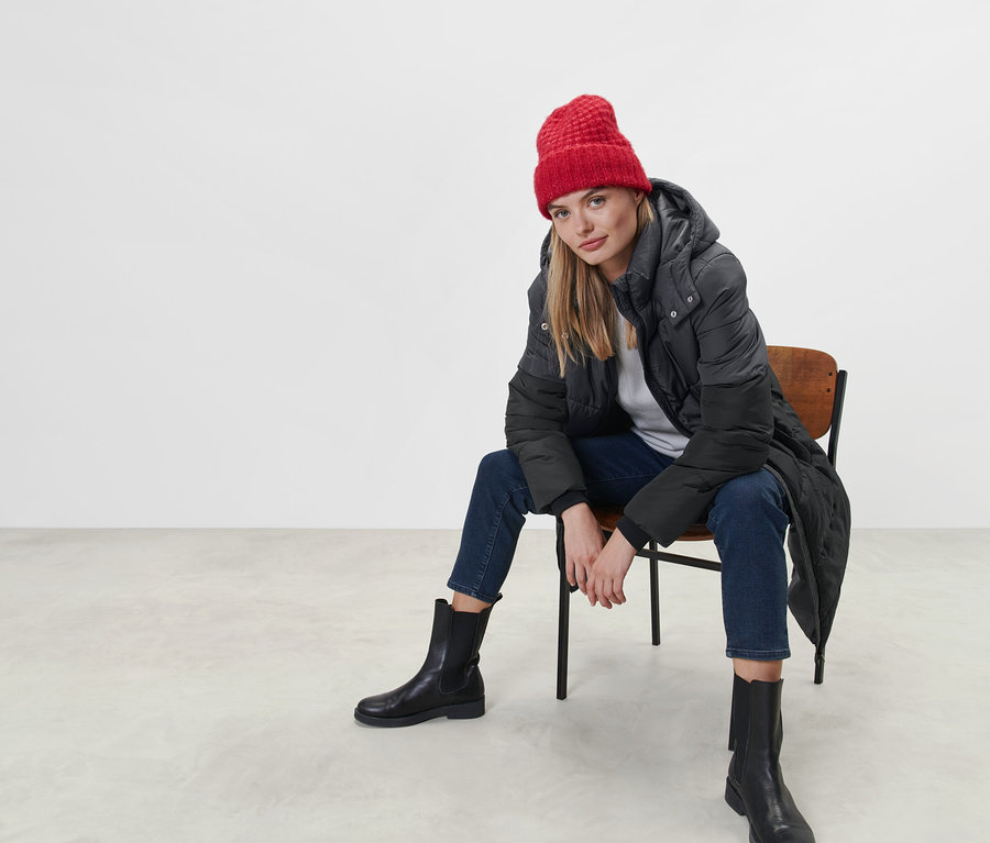 Nő ül egy széken, steppelt kapucnis kabátot és piros sapkát visel.