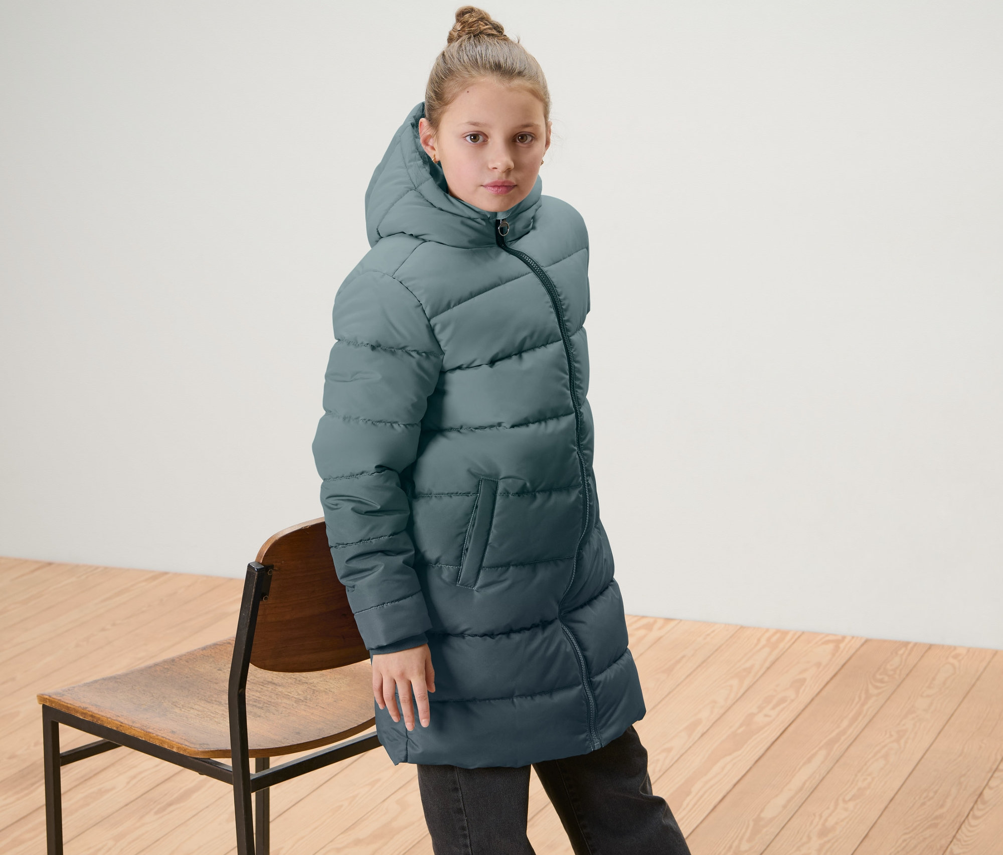 Egy lány színátmenetes gyerek termokabátot visel, és egy fa szék mellett áll.