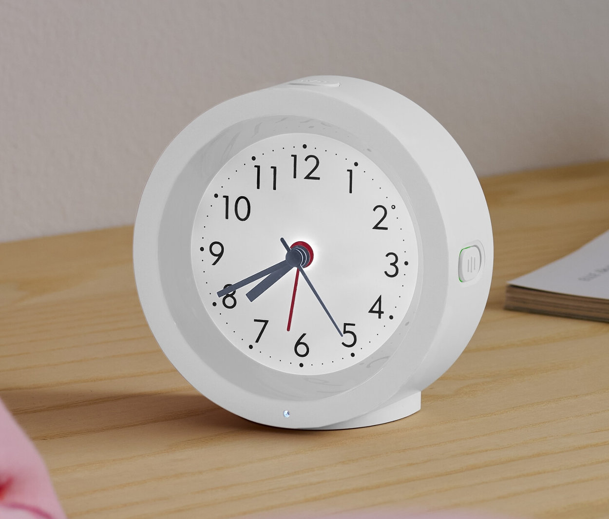 Egy fehér analóg kvarc ébresztőóra háttérvilágítással egy éjjeliszekrényen áll egy ágy mellett.