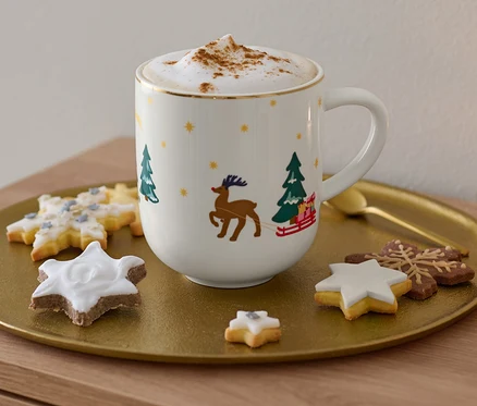 Egy arany tálcán egy nagyméretű, karácsonyi mintával díszített bögre, egy cappuccino és sütemények láthatók.
