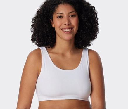 Mosolygó, göndör hajú nő fehér SCHIESSER bustier melltartót és fehér alsóneműt visel, egy fehér tömbnek támaszkodva.
