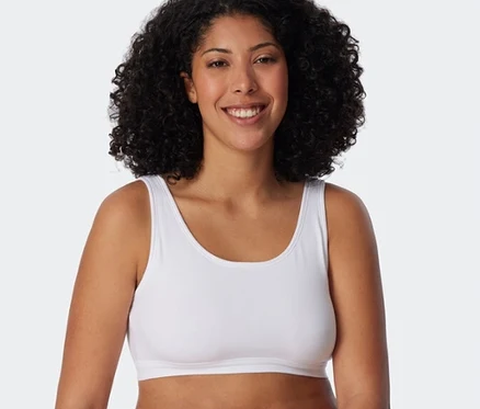 Mosolygó, göndör hajú nő fehér SCHIESSER bustier melltartót és fehér alsóneműt visel, egy fehér tömbnek támaszkodva.