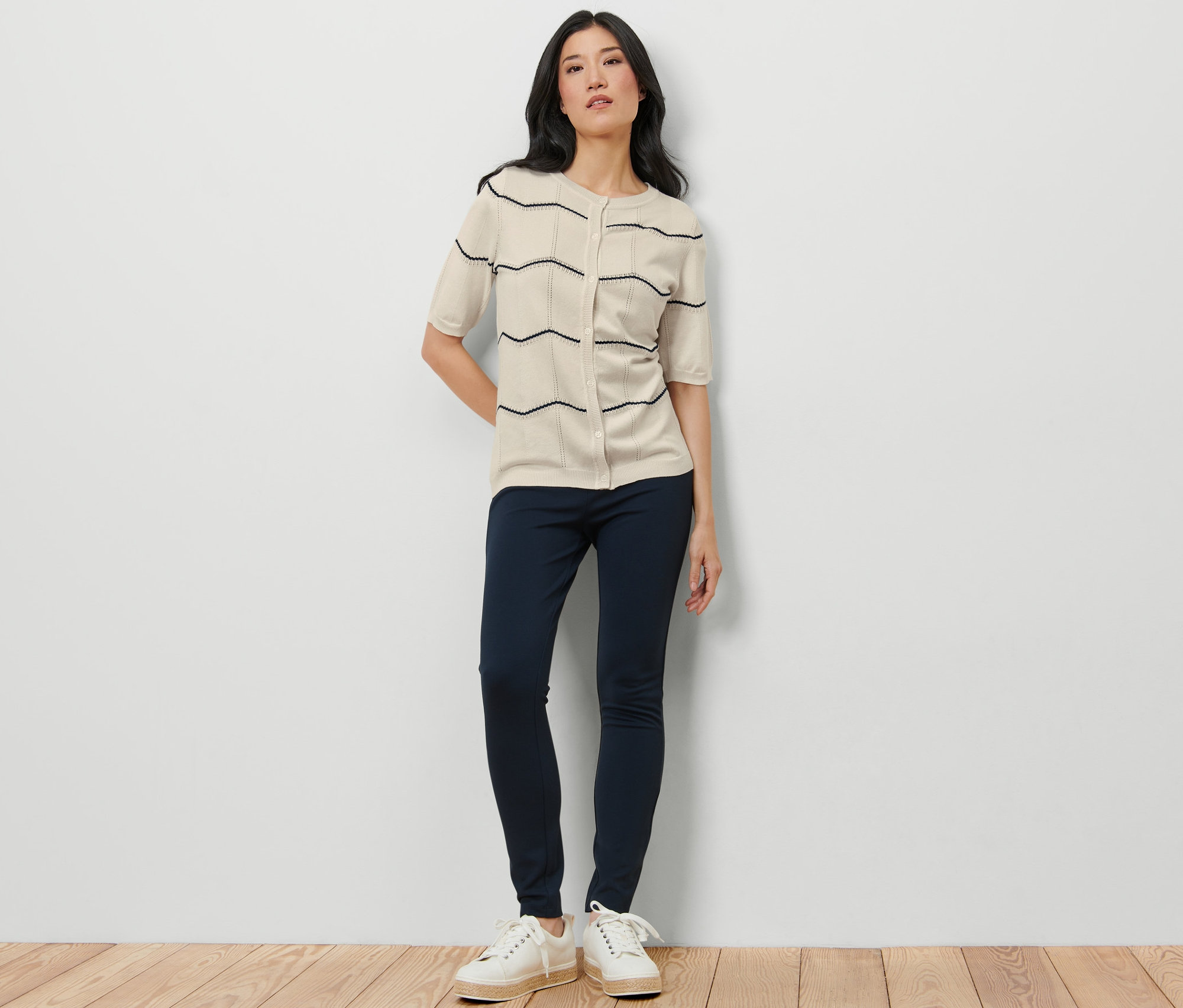Egy nő áll, és azsúrmintás finomkötésű kardigánt, sötétkék nadrágot és fehér tornacipőt visel.
