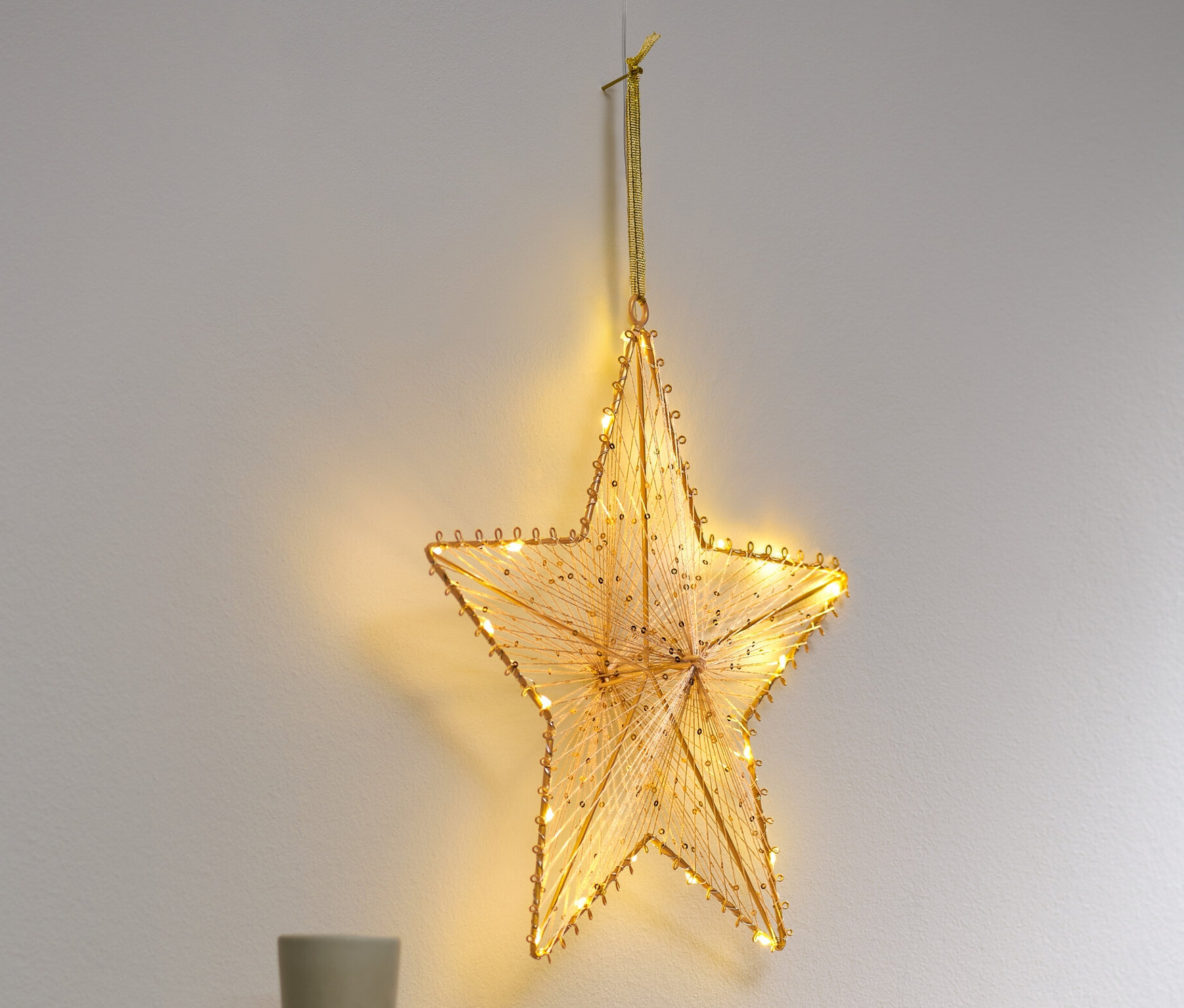 Egy LED-es csillag függődísz lóg a falon egy szekrény felett, amelyen váza és könyvek találhatók.