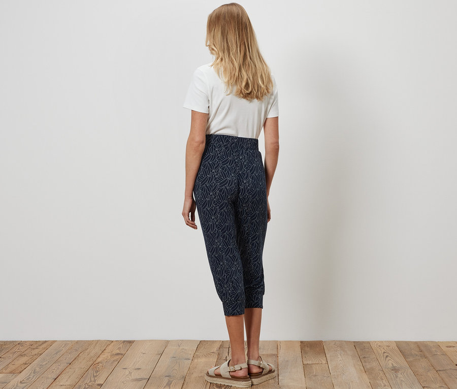 Hátulról látható nő áll a fapadlón, kék-krémfehér 3/4-es jersey nadrágot és fehér pólót visel.