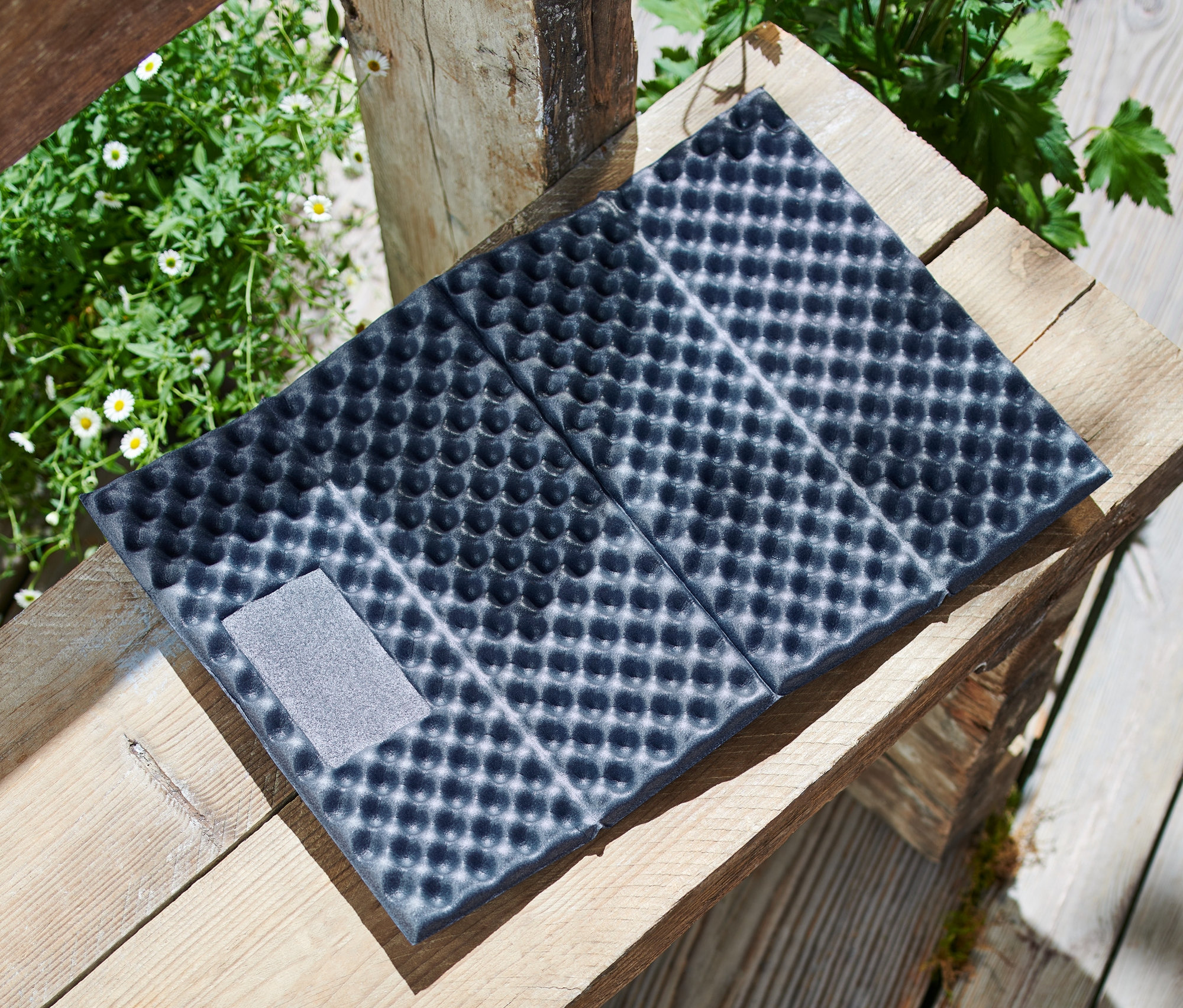 Egy összehajtható termo ülőpárna fekszik egy fa raklapon virágok mellett.