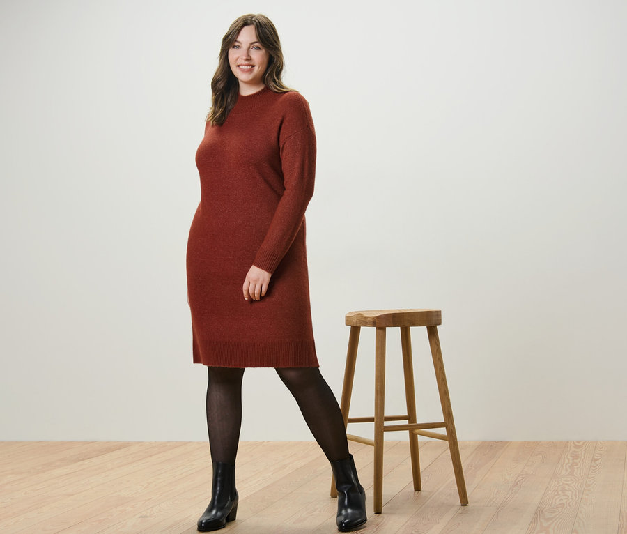 Nő pózol kötött ruhában, gyapjúval és 50 DEN-es, átlátszó hatású harisnyában egy fa szék mellett.