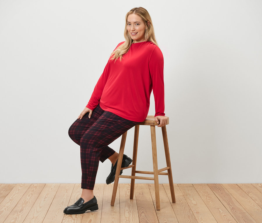 Egy nő ül egy széken, kockás sztreccsnadrágot és fodros blúzt visel.