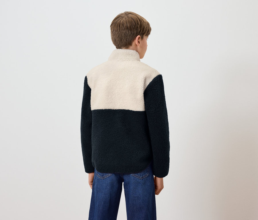 Egy fiú háttal a kamerának kék farmerban és egy gyapjú pulóverben, krém színű felsőrésszel és sötétkék aljával.