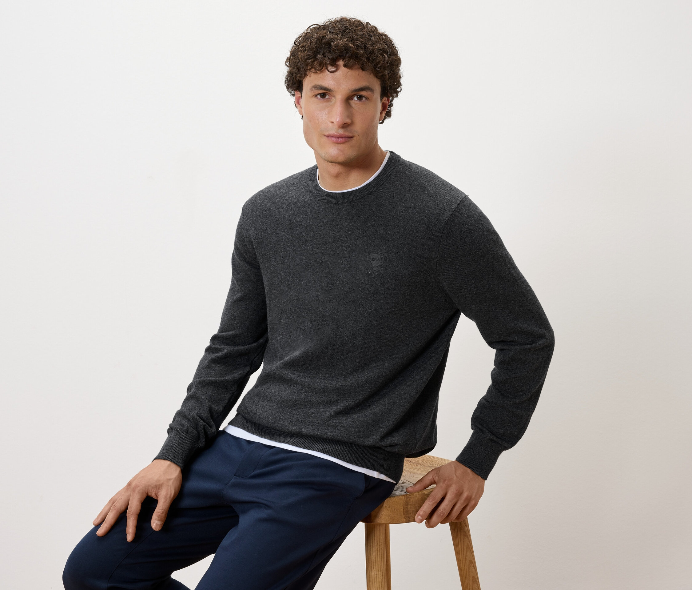 Egy férfi ül egy széken, szürke pulóvert és sötétkék nadrágot visel.