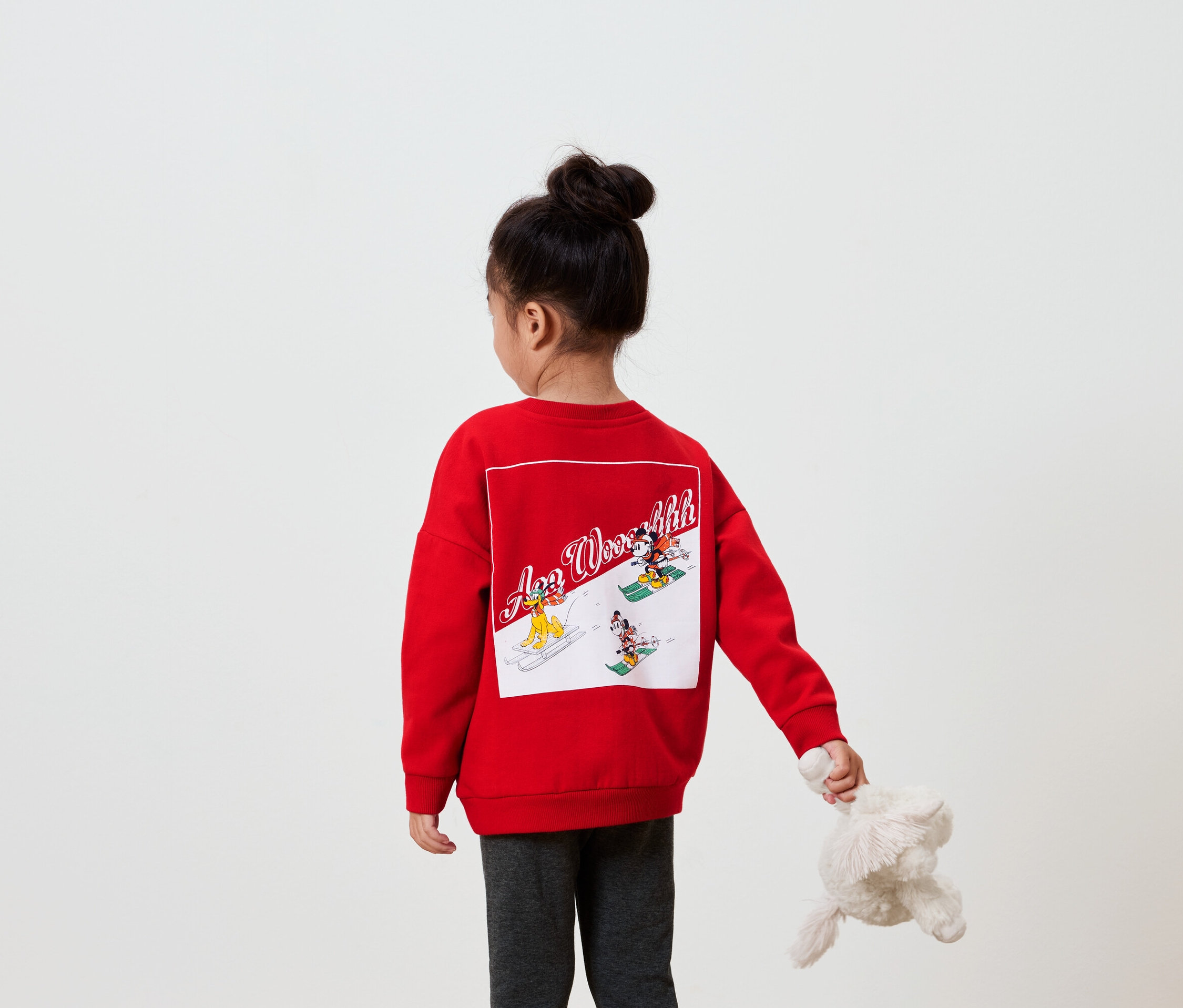 A fényképezőgépnek háttal álló lány piros pulóvert visel, hátulján képpel, és egy plüssállatot tart a kezében.