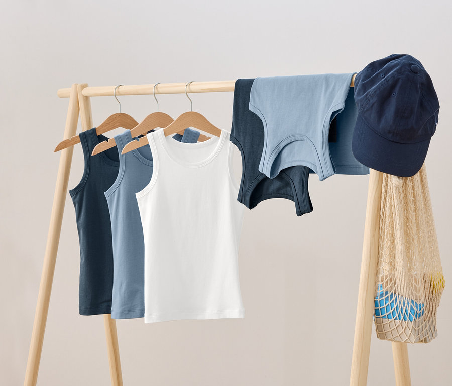 Egy fa vállfán három trikó lóg vállfán, mellette két másik trikó, egy kék baseball sapka és egy hálós táska.