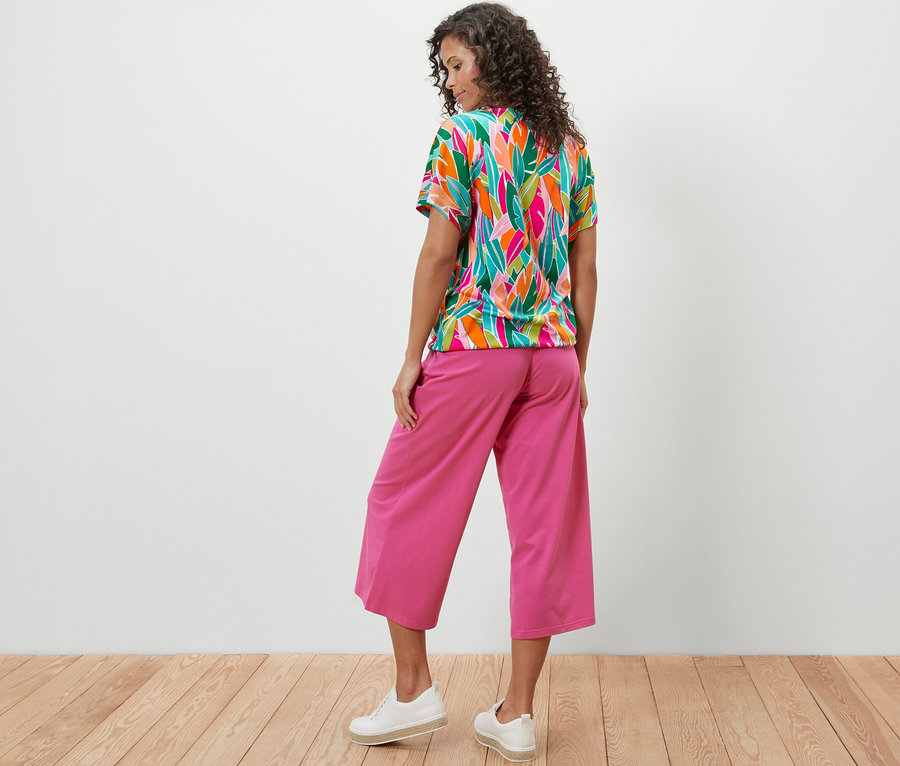 Hátulról látható nő ejtett vállú, nyomott mintás pólóblúzban és rózsaszín nadrágban.