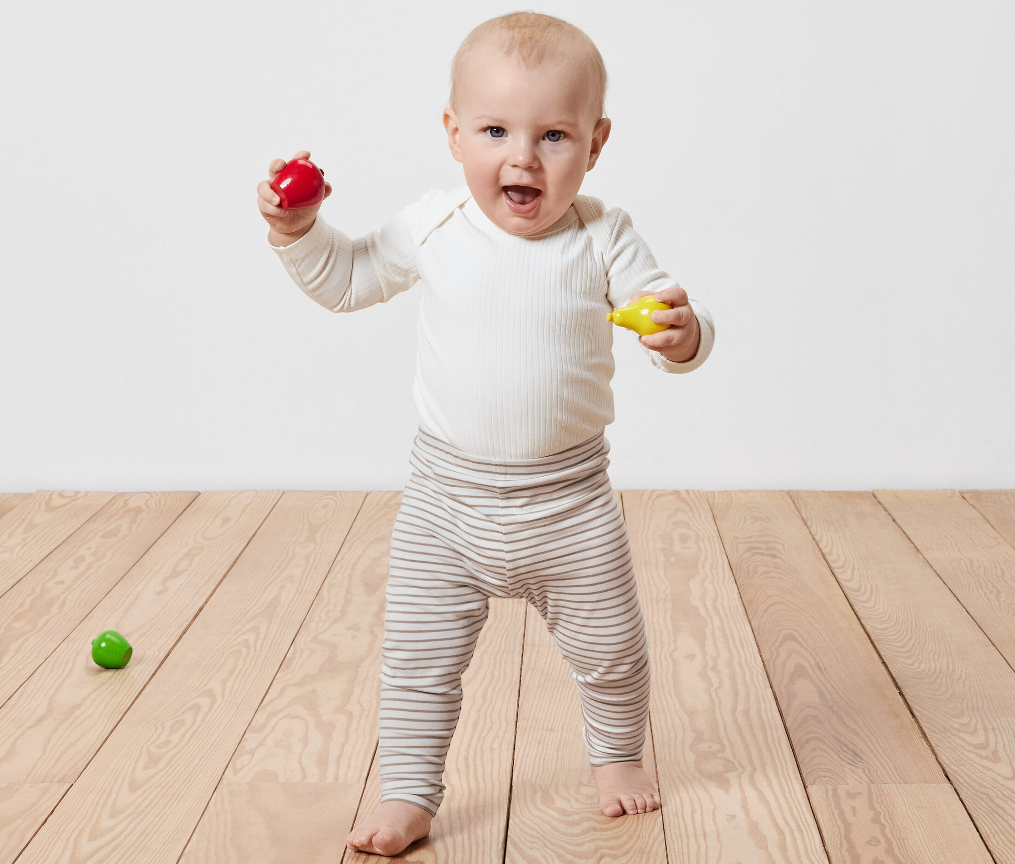 Egy mosolygó baba áll a fapadlón, kezében piros és sárga játékot tart. 5 gyerekleggings van rajta.