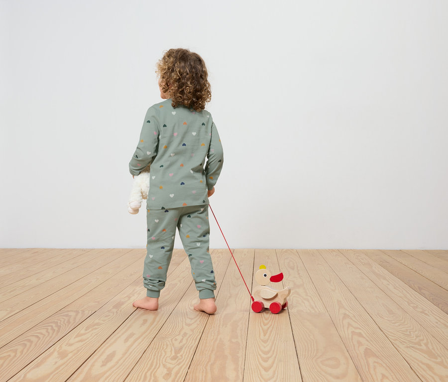 Egy szívmintás pizsamában lévő gyermek egy kerekes fa kacsa játékot húz maga után, és egy plüssmackót tart a kezében.