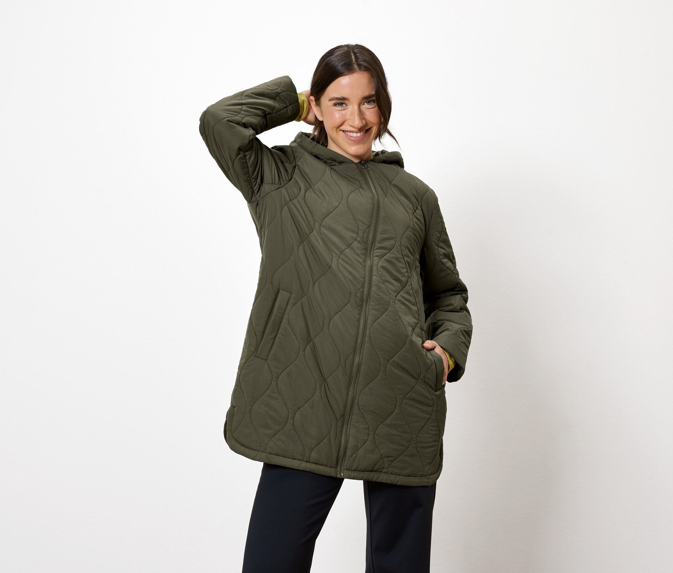 Nő pózol olívazöld, steppelt, kapucnis kabátban és sötétkék nadrágban.